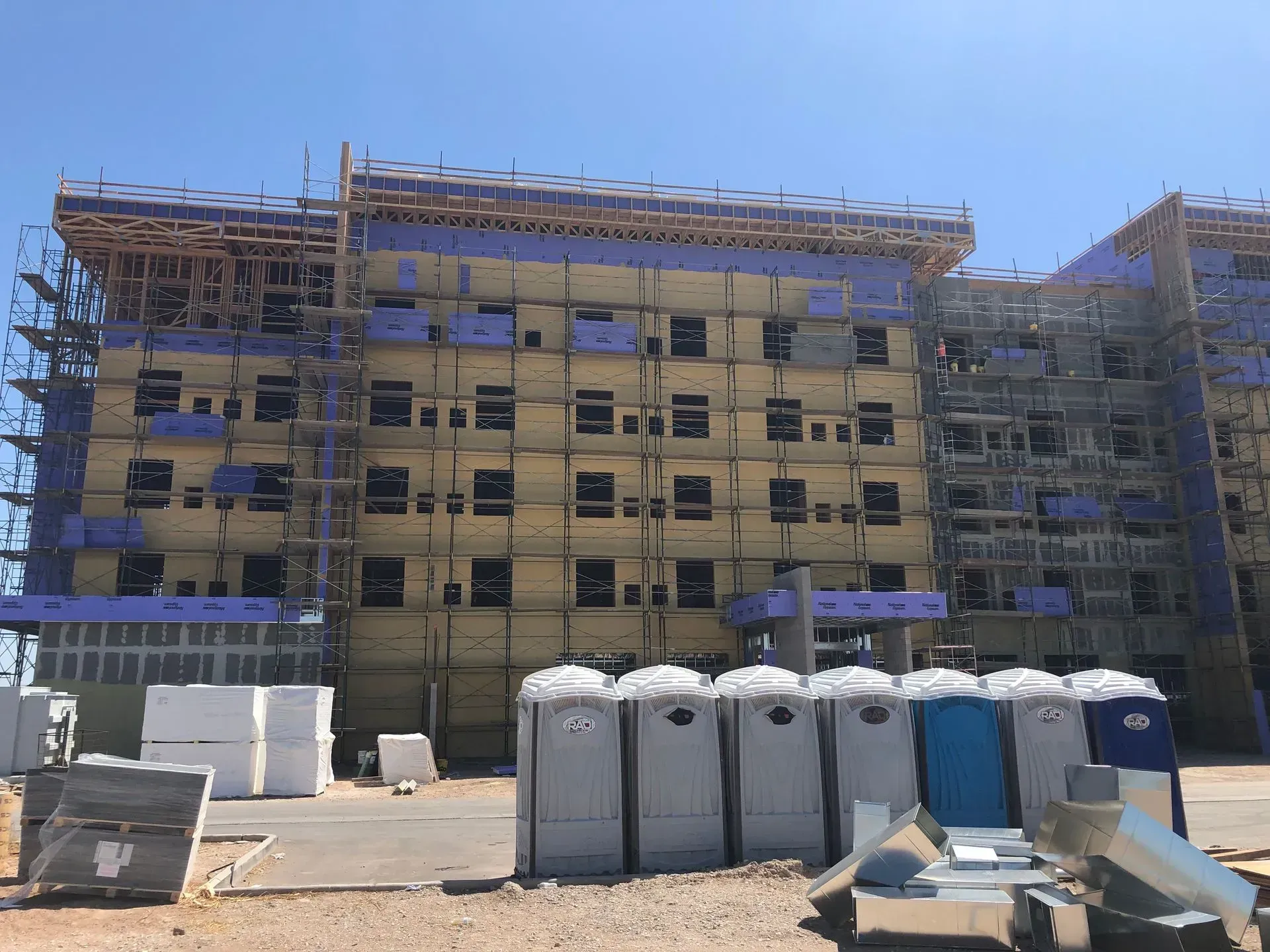 Construction site with a multi-story building under scaffolding and portable toilets in front.