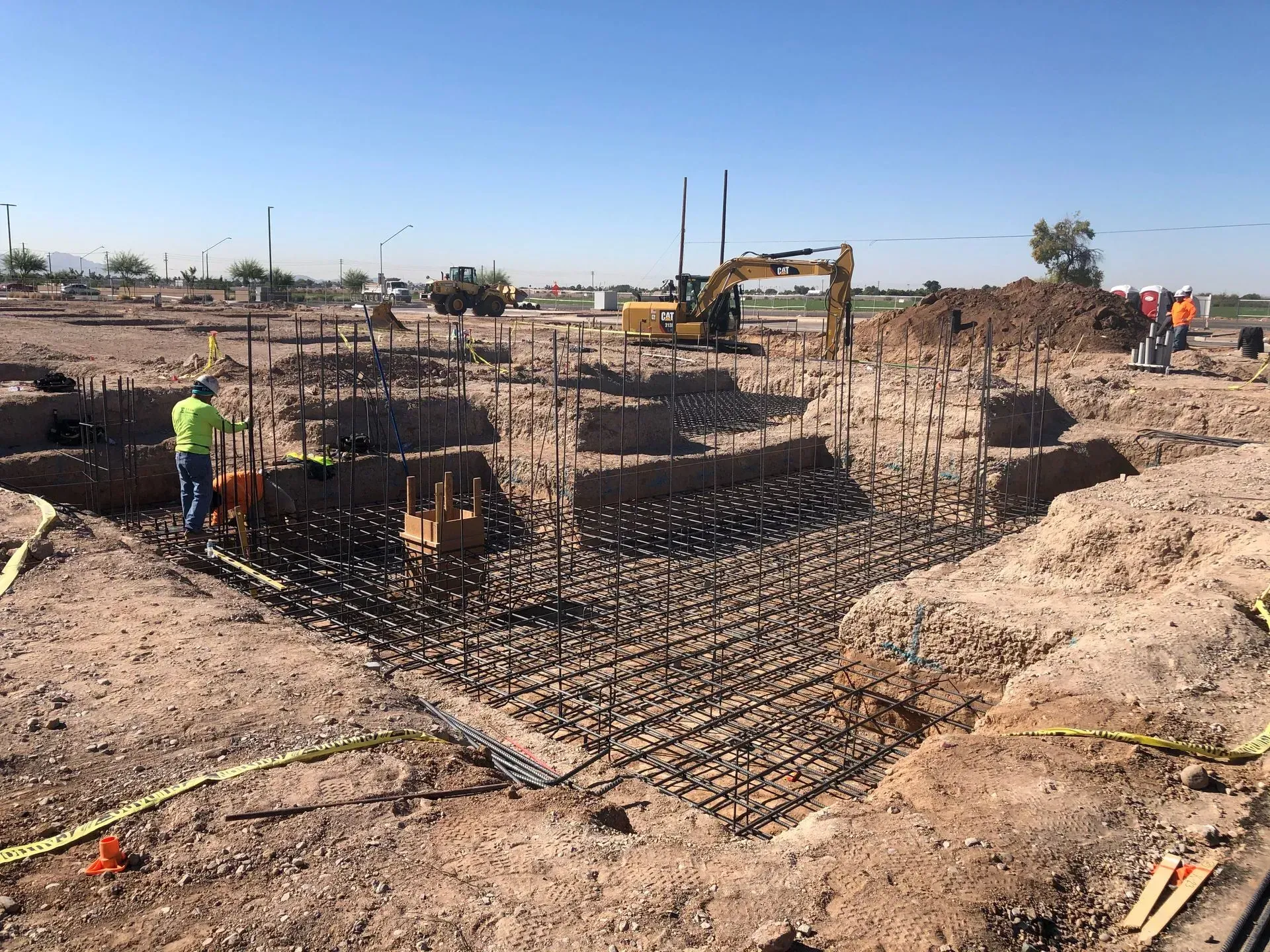 Construction site with rebar foundation trenches and an excavator in a dusty field under a clear sky