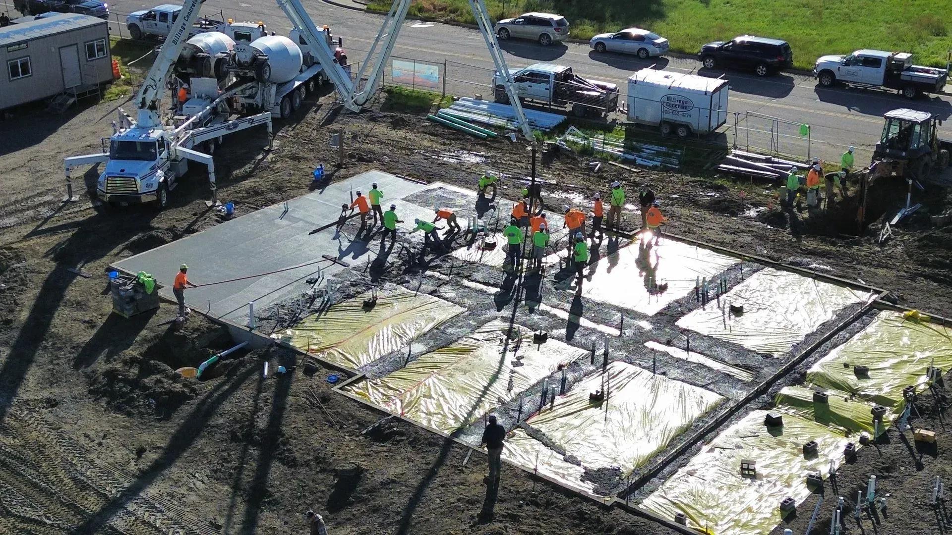 Construction crew pouring concrete on a foundation slab beside a blue pump truck and parked vehicles.