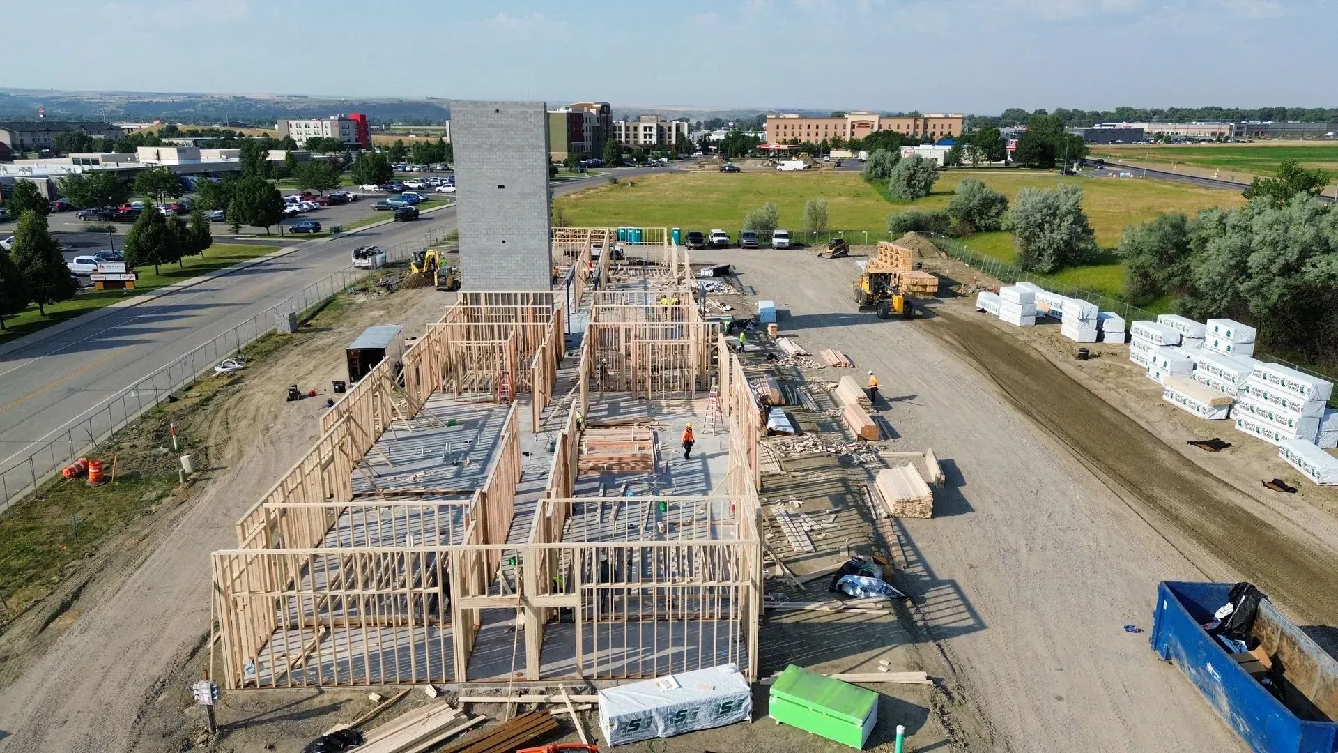 Aerial view of a wooden house under construction beside a road, with machinery and stacked building materials nearby.
