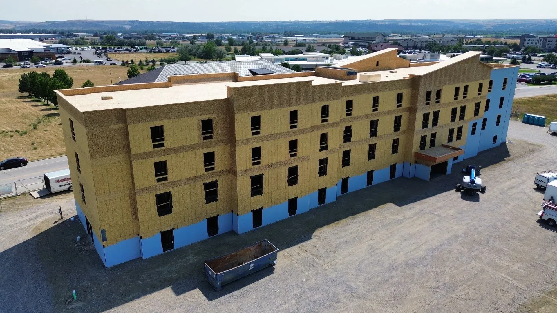 Partially finished tan multi-story apartment building with blue lower panels on a gravel lot under clear skies