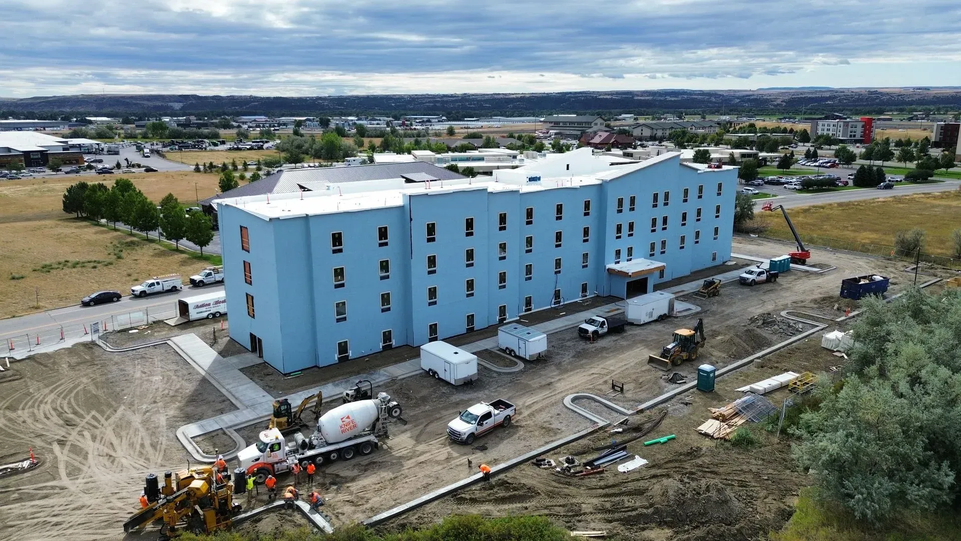 Blue multi-story apartment building under construction with workers, trucks, and equipment on a dirt site.