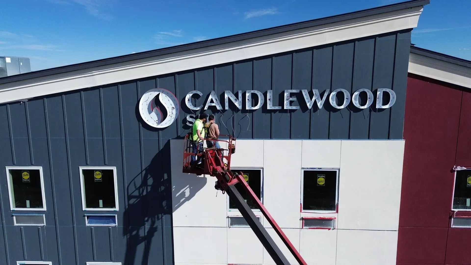 Workers on a ladder installing the Candlewood sign on a gray and maroon building under a blue sky