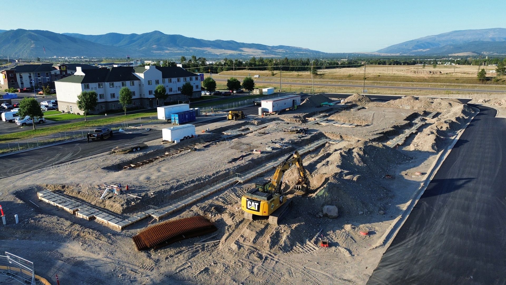 Construction site with excavator, trenches, and scattered materials beside a road, mountains in the distance