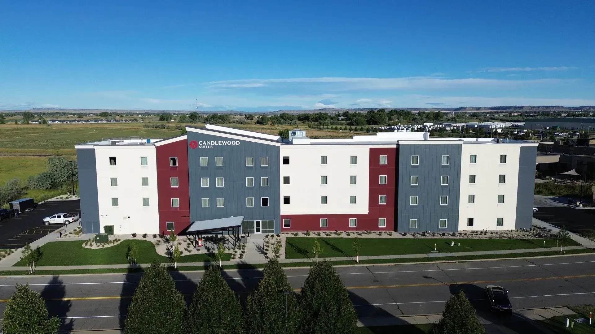 Aerial view of a modern apartment building with white, gray, and maroon panels beside a road and trees
