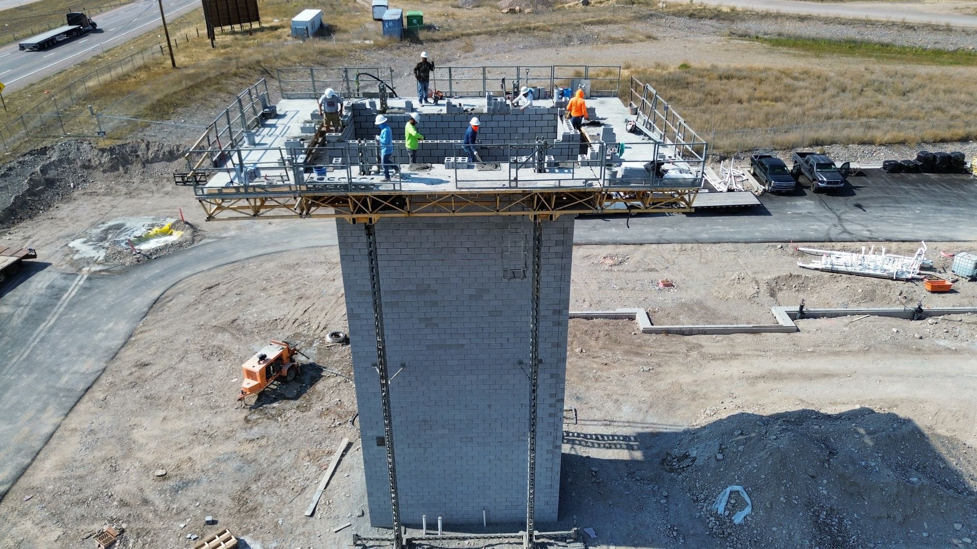 Construction crew atop a concrete bridge pier on a worksite, with equipment and vehicles nearby.
