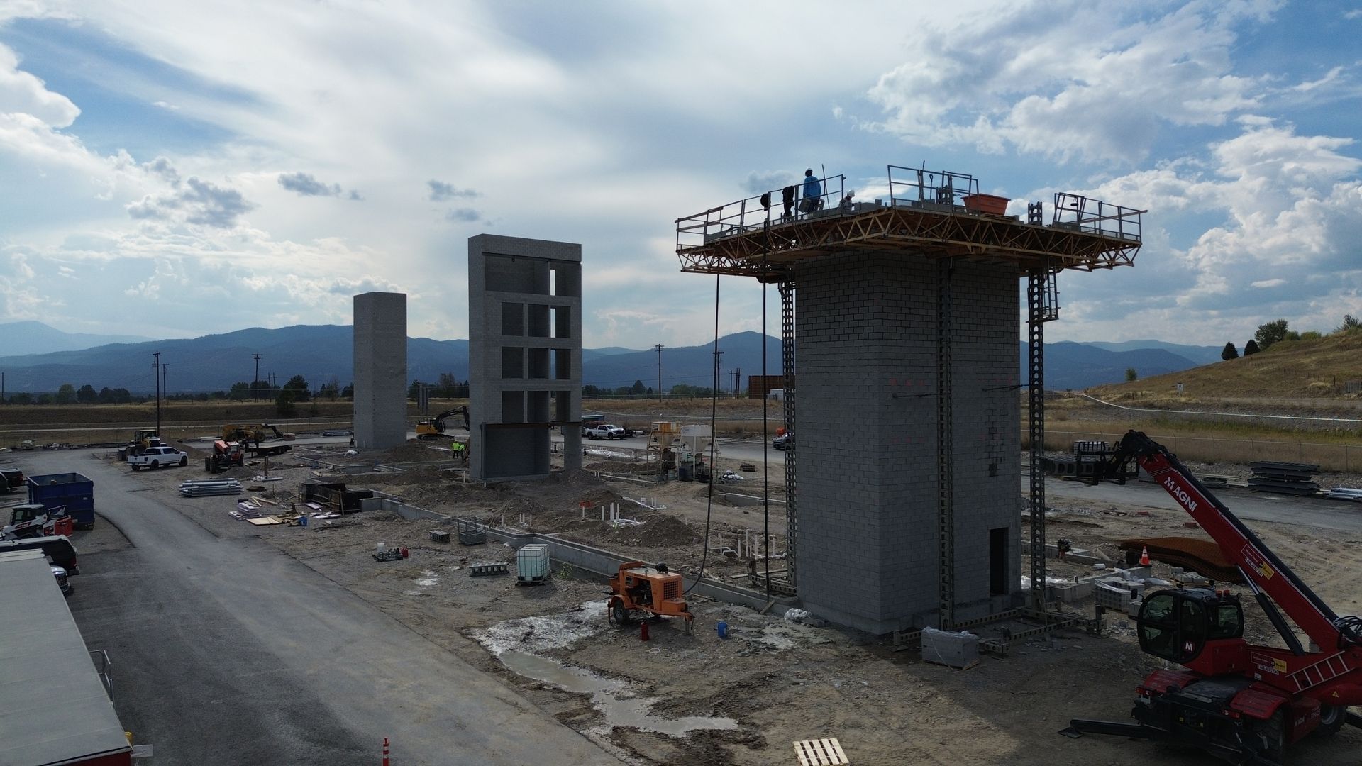 Construction site with two tall concrete bridge piers and heavy machinery beside a road under a cloudy sky