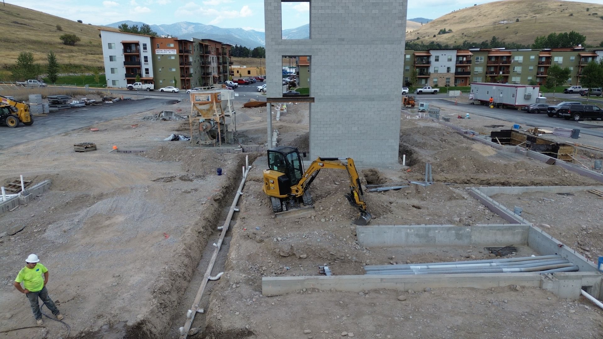 Construction site with excavator beside a concrete tower and foundation trenches.