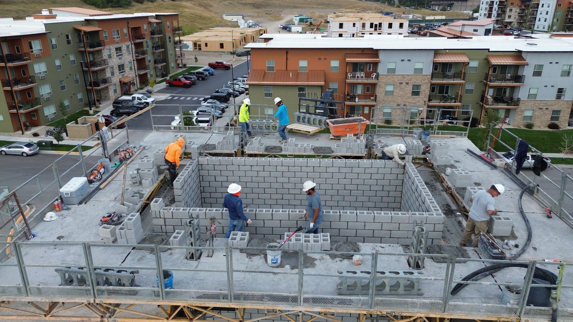 Construction workers on a rooftop wall at an apartment complex, with tools and city buildings in the background.