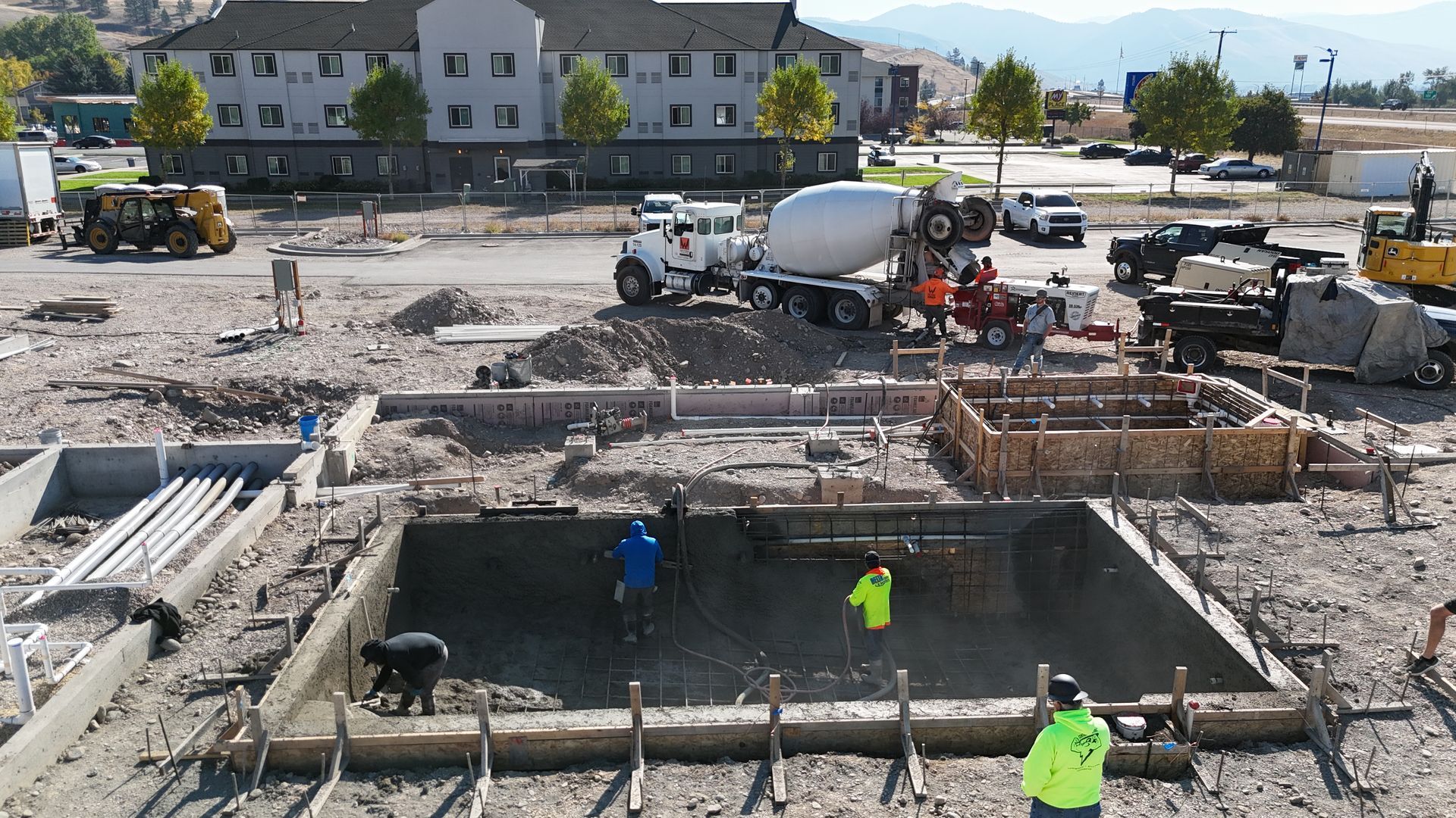 Construction site with workers, concrete forms, and excavators around a large rectangular foundation pit.
