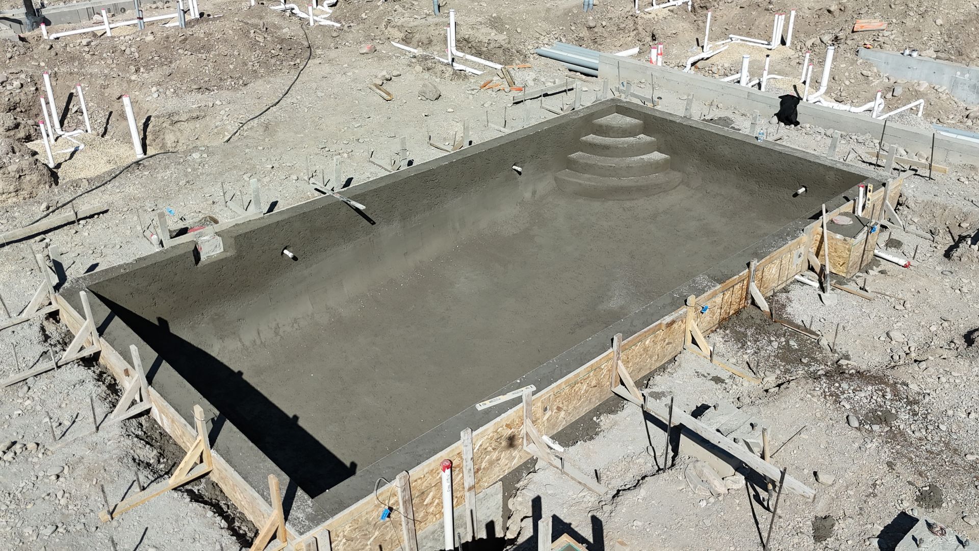 Concrete foundation slab under construction in a dirt excavation site, with wooden forms and rebar stakes nearby