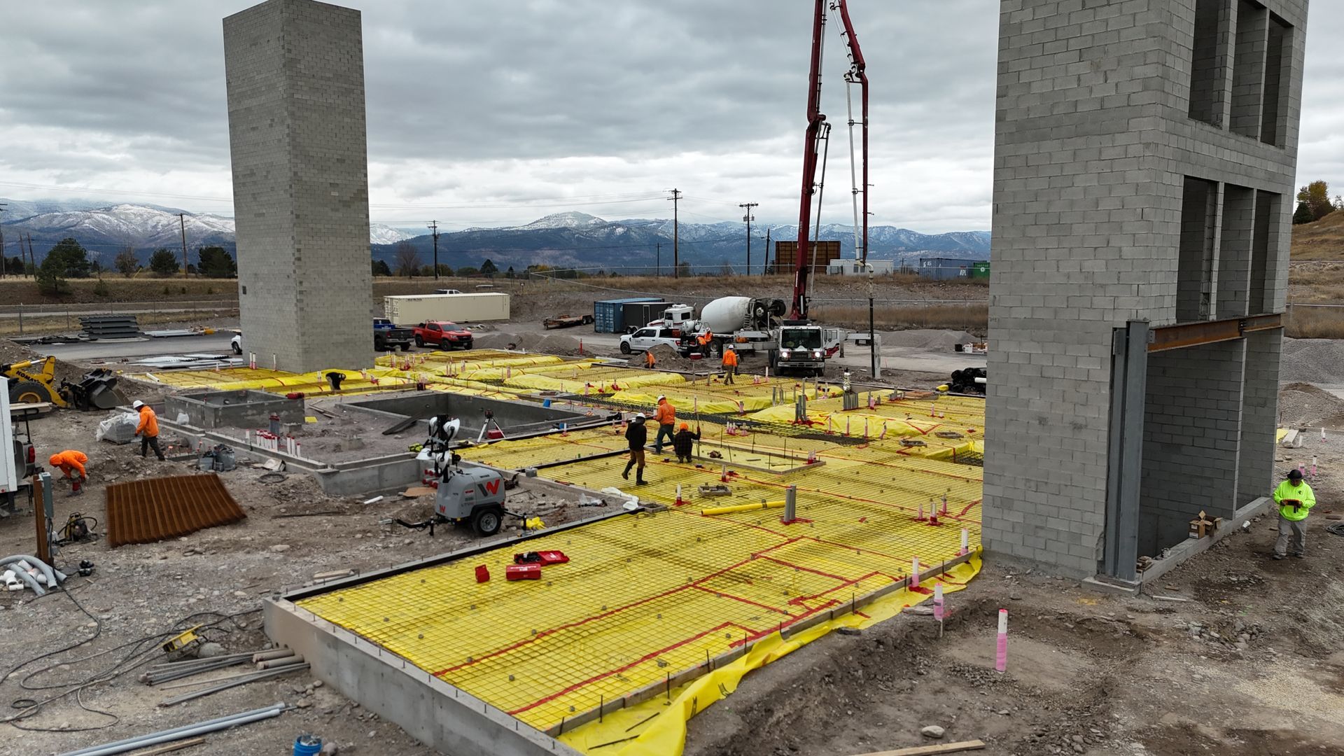 Construction site with workers on yellow floor panels beside concrete walls and a crane under overcast sky
