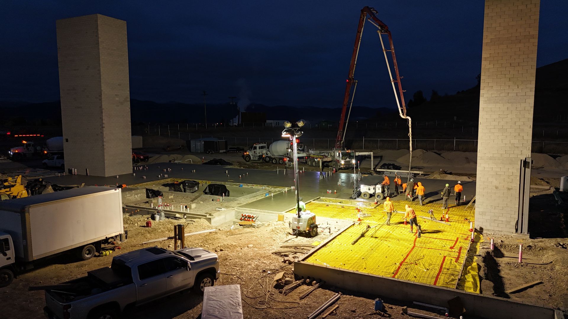 Nighttime construction site with cranes, workers, trucks, and a large yellow-lit slab area.