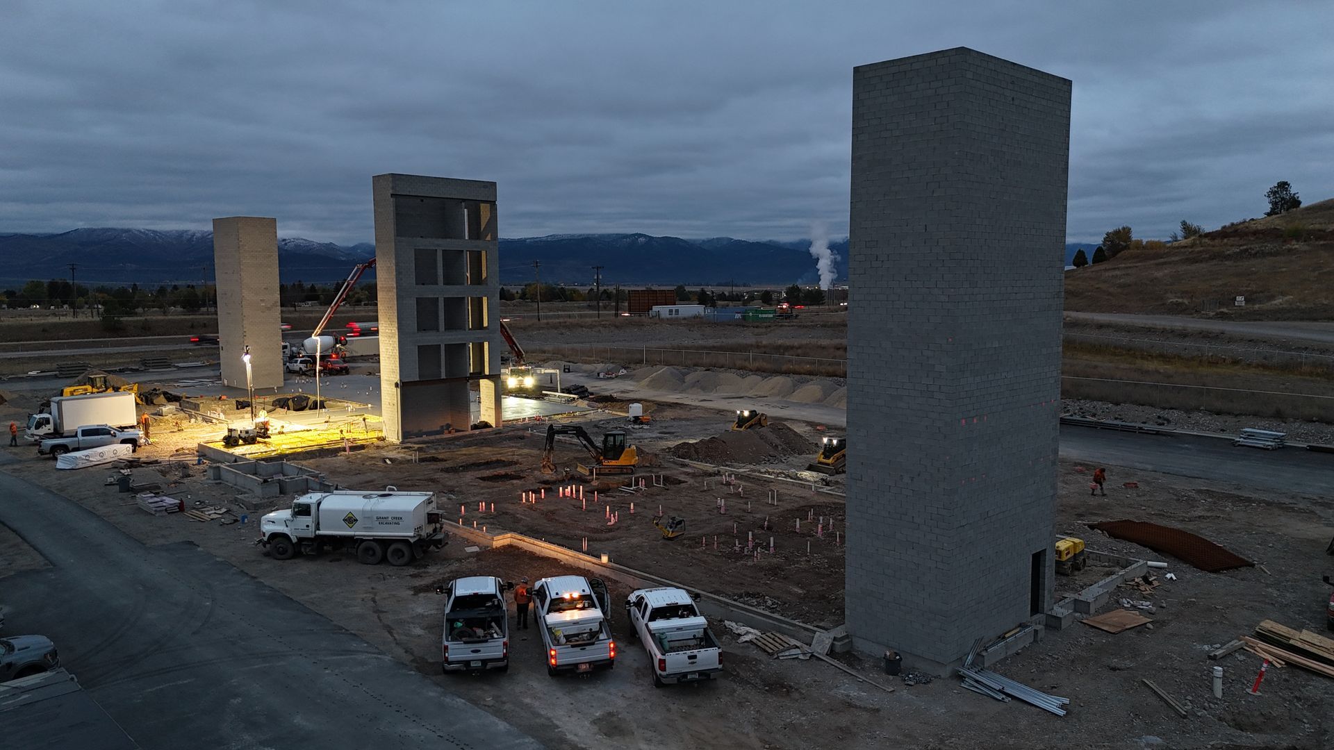 Construction site with two tall concrete structures, work lights, vehicles, and workers at dusk