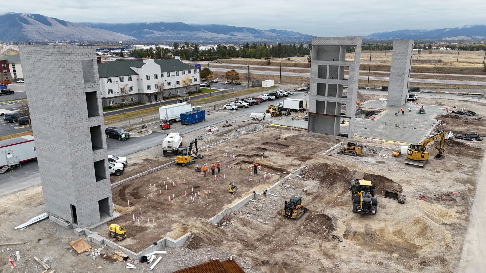 Construction site with multiple concrete towers, heavy equipment, and workers on a muddy lot.