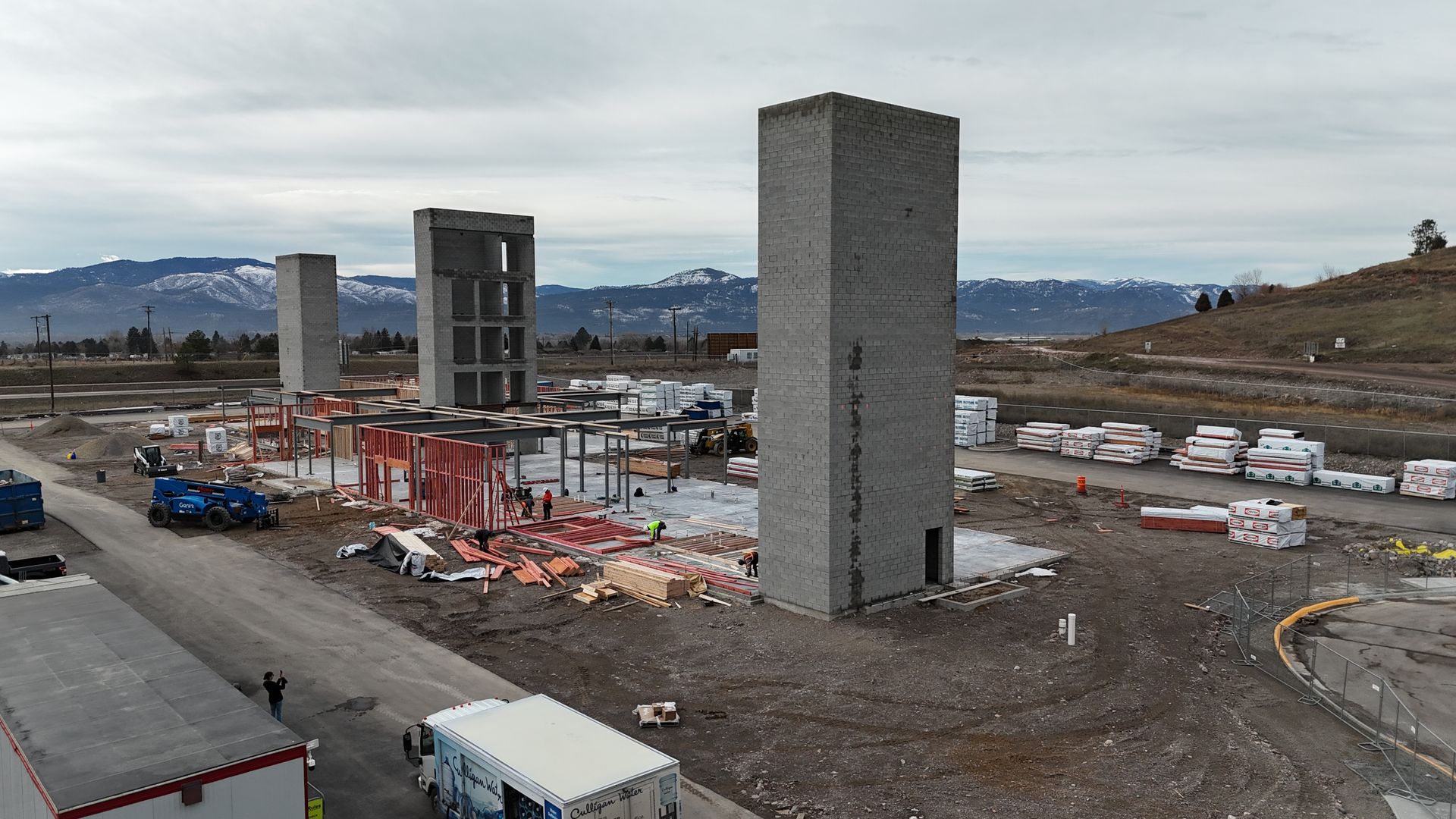 Construction site with two tall concrete towers, cranes, and scattered materials on a gravel lot by the water.