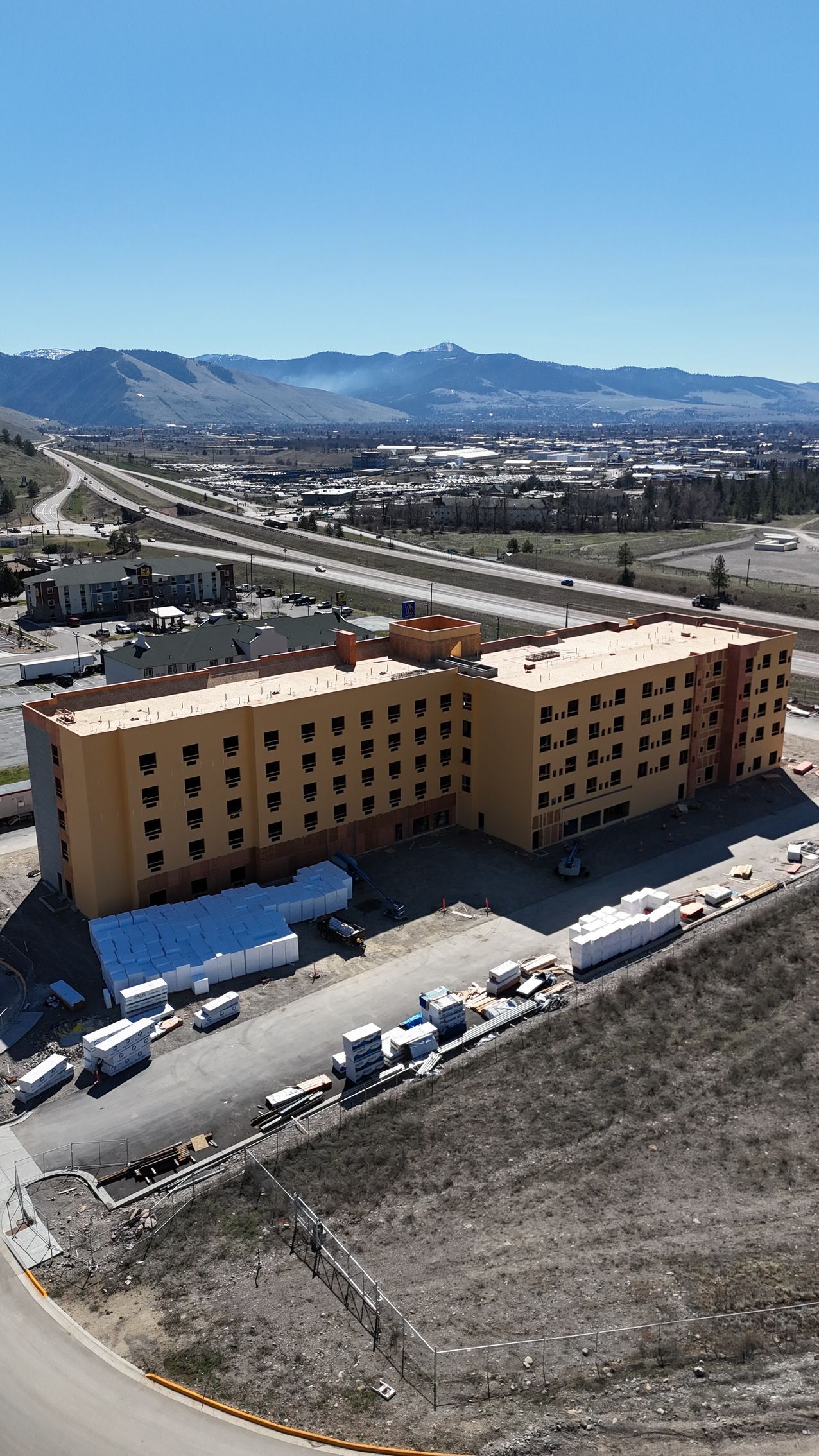 Yellow apartment building beside a road, with snowy hills and mountains under a clear blue sky.