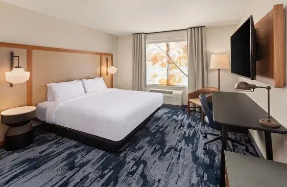 Modern hotel room with a large bed, desk, wall-mounted TV, and window view of autumn trees