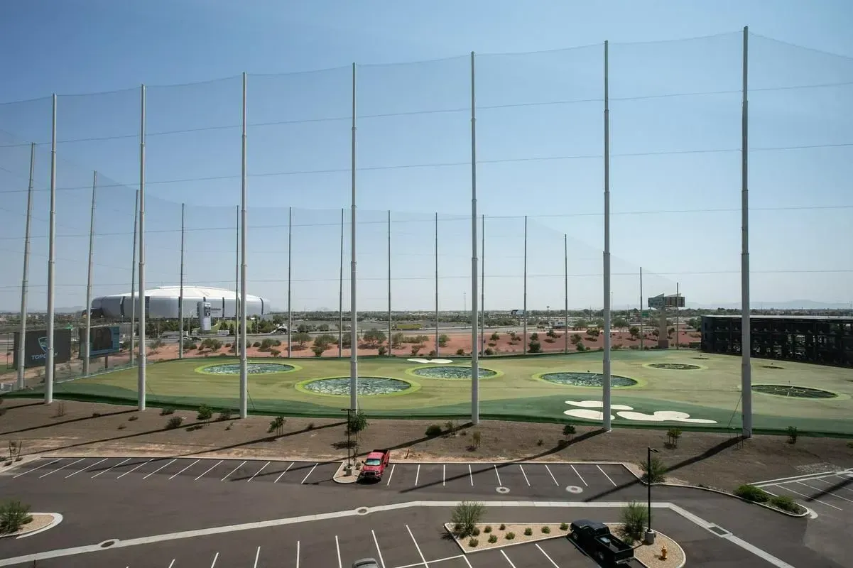 Baseball practice field with tall netting, green turf, and parked cars under a clear blue sky