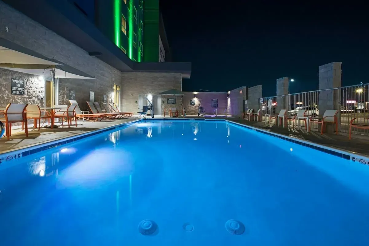 Rooftop pool at night with blue-lit water, lounge chairs, and city skyline lights in the background