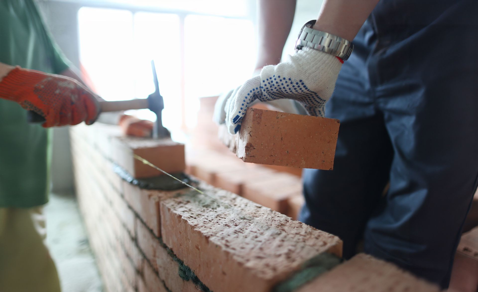 Two construction workers in gloves carefully laying red bricks to build a wall.