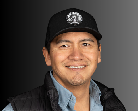 Man wearing a black hat smiles at the camera, wearing a blue shirt and black vest; neutral gradient background.