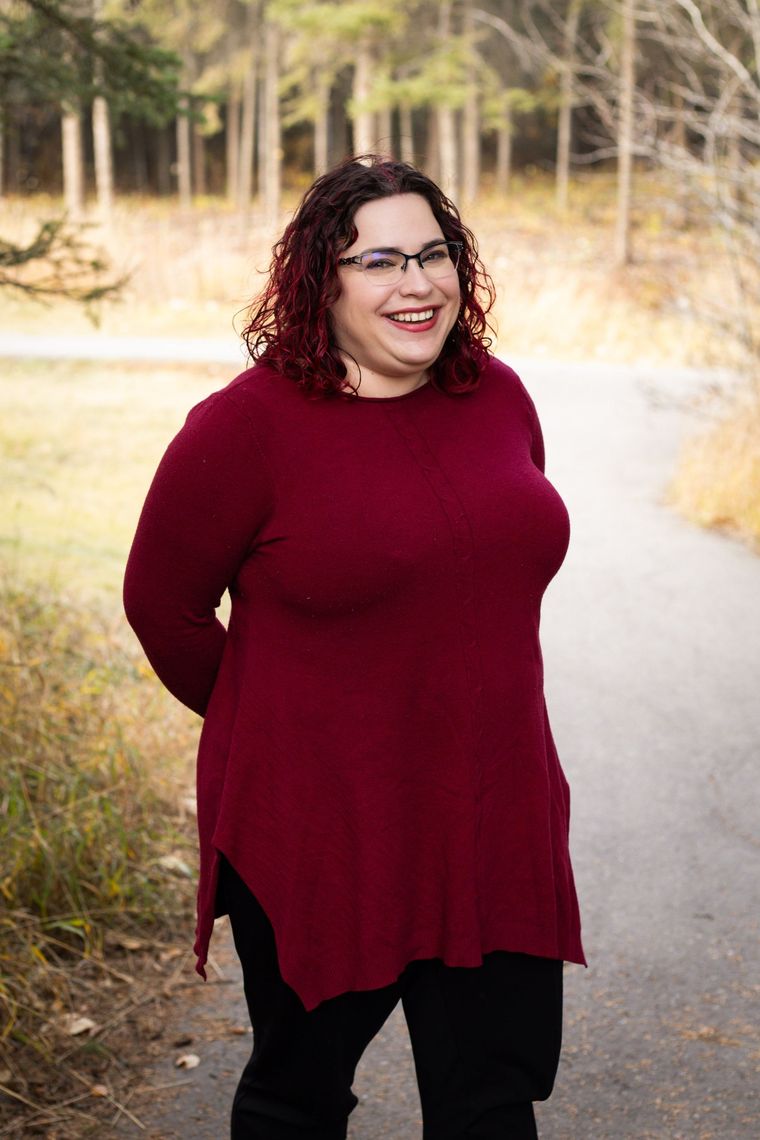 Woman smiles, standing on a path in a park; wearing a red sweater and glasses.