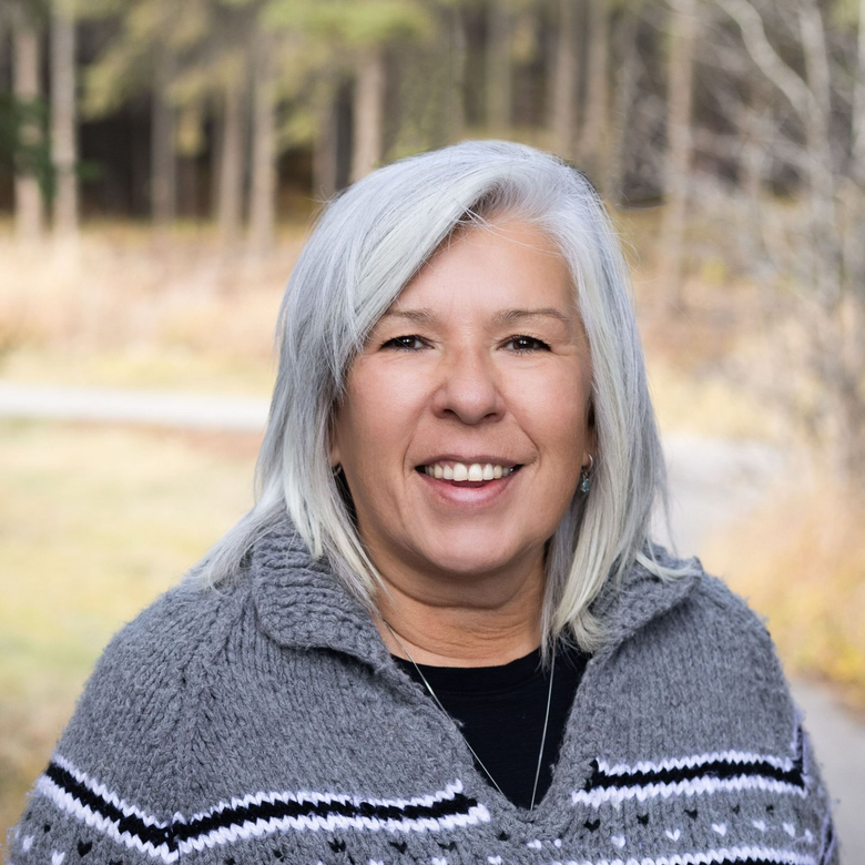 Woman with gray hair smiles, wearing a gray and white patterned sweater; outdoor background.