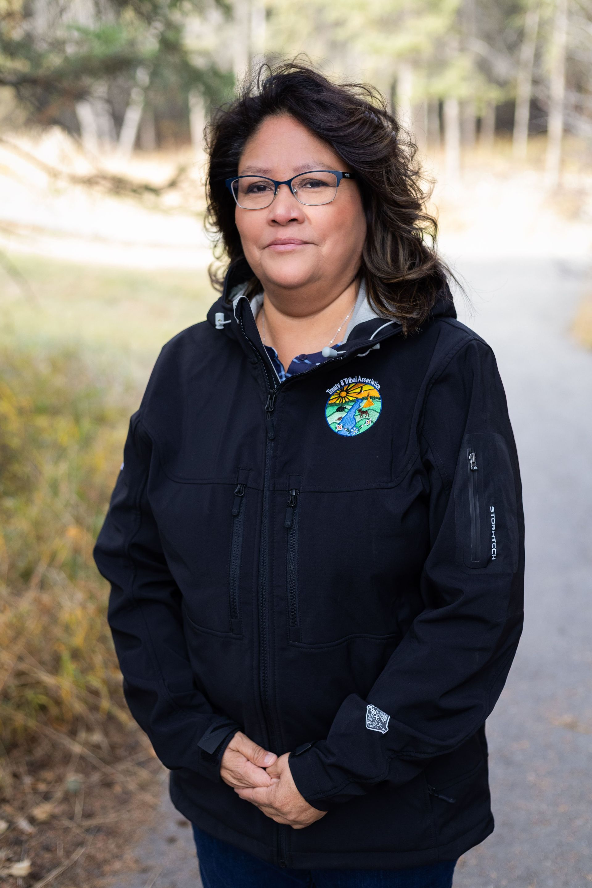 Woman in black jacket with a patch, standing on a path in a wooded area.