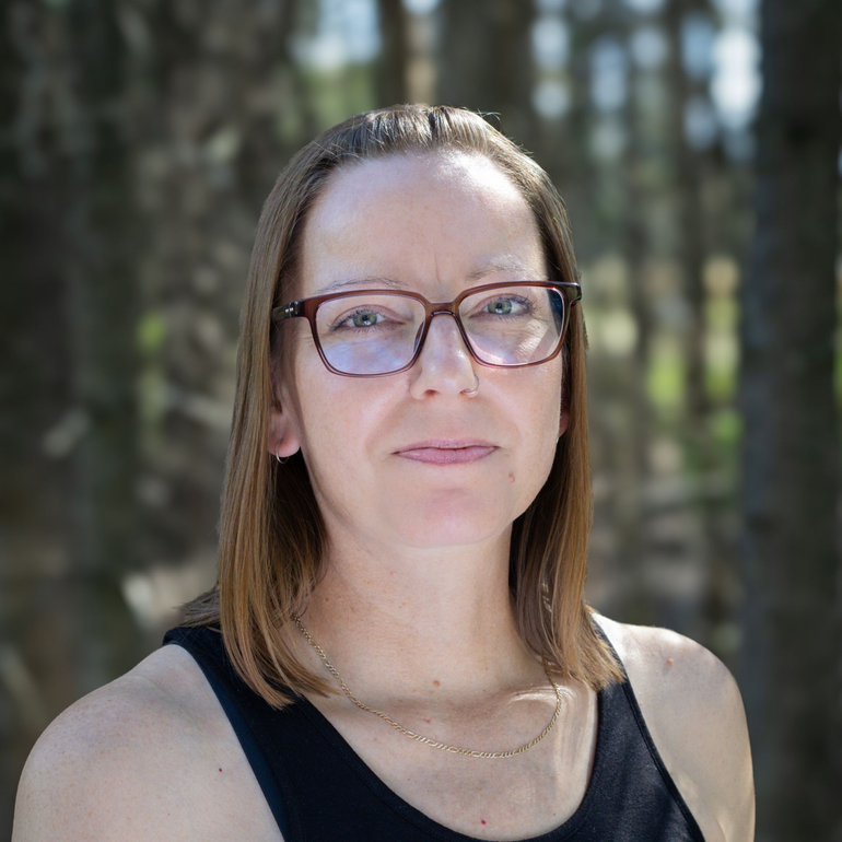 Woman with graying hair wearing a black and white heart-patterned shirt, standing on a path in a forest.