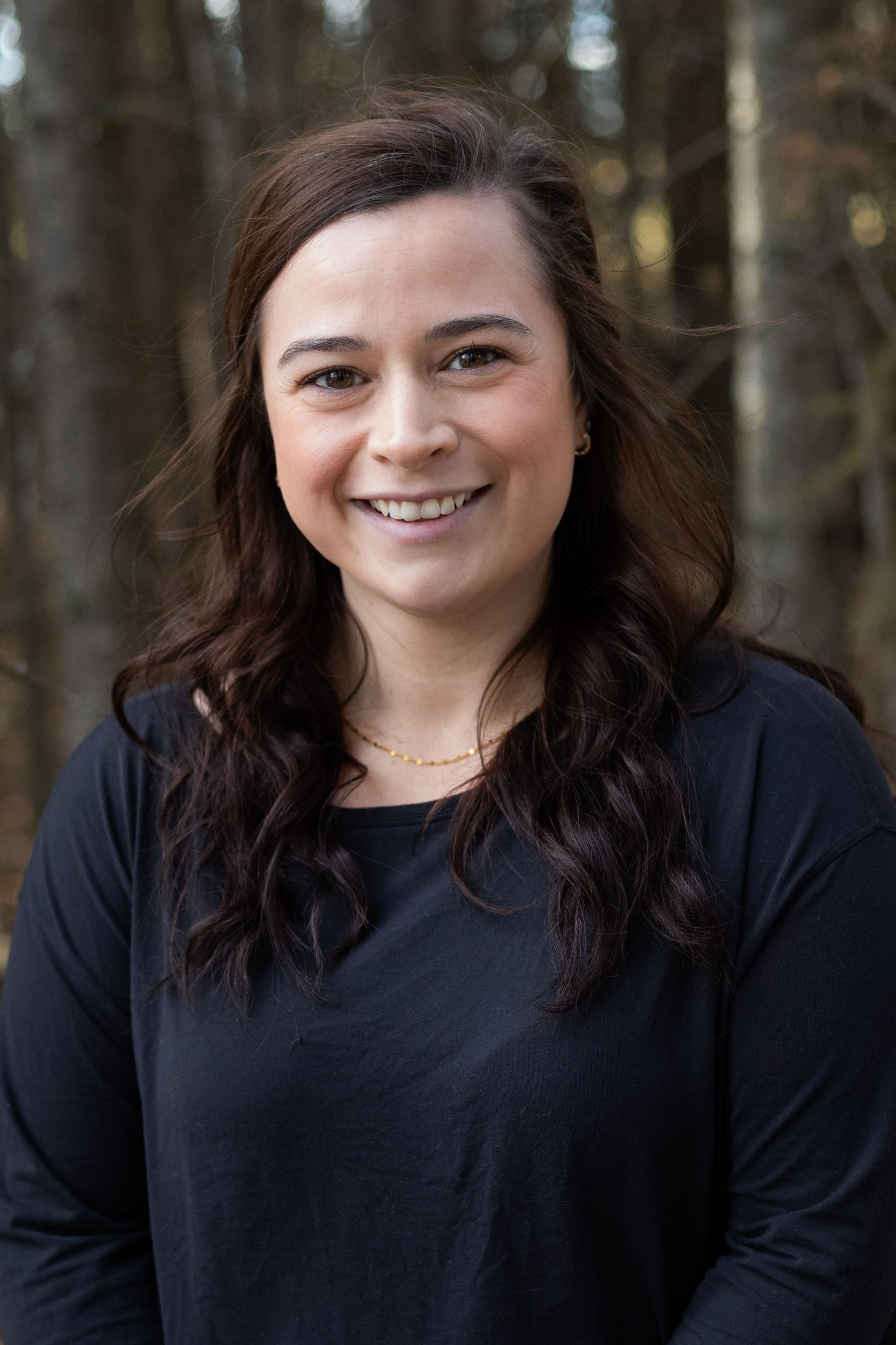 Woman smiling in front of blurred trees, wearing a black top and gold necklace.
