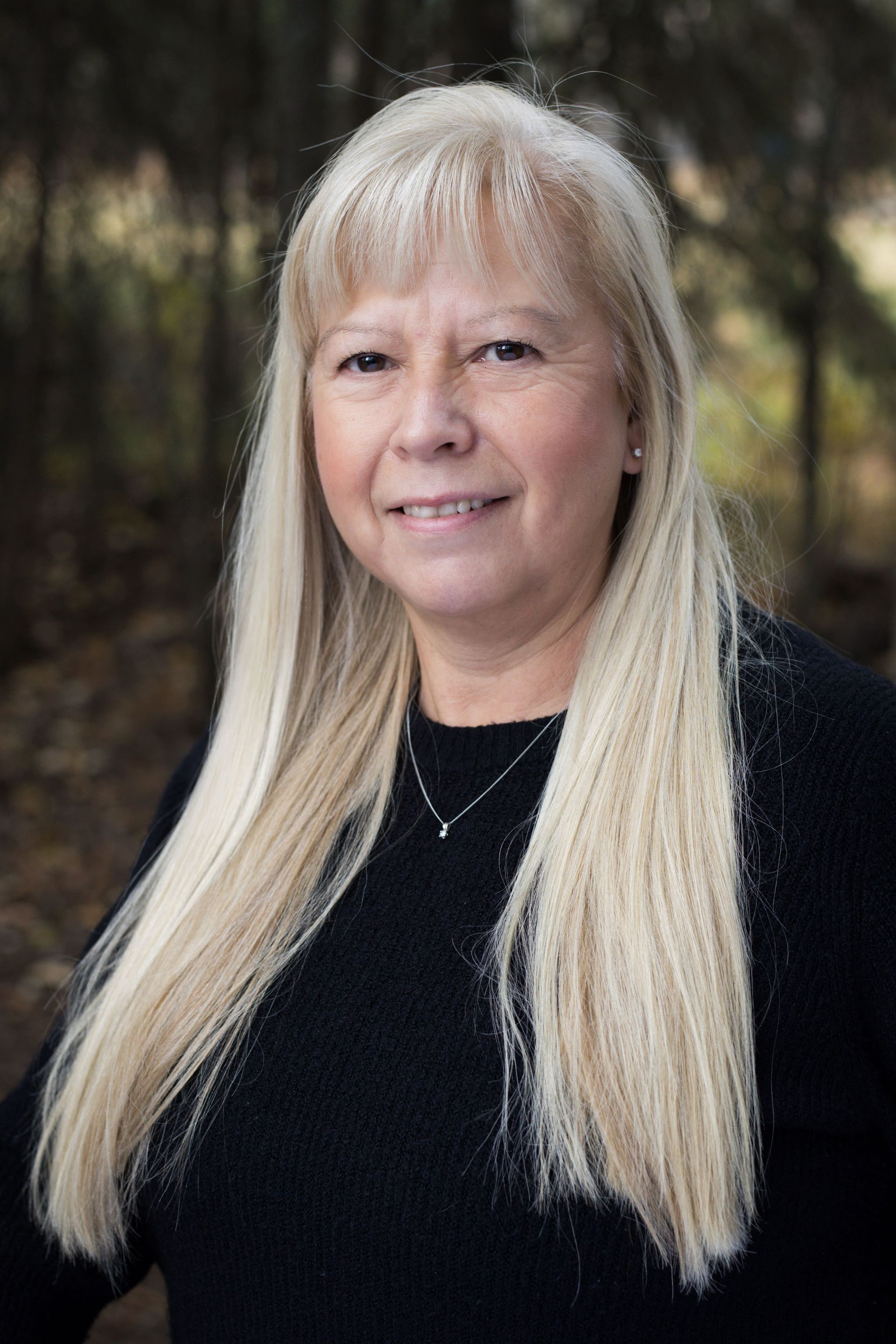 Woman with long blonde hair, smiling, wearing a black sweater, set in a forest.