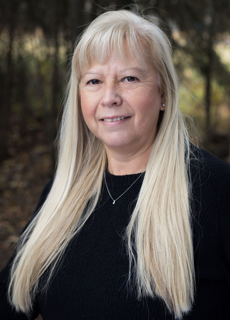 Woman with long blonde hair, smiling; wearing black sweater; outdoors with trees.