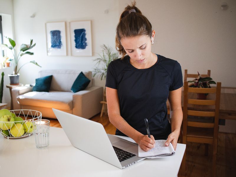 Frau am Laptop im Call mit Headset, schreibend im Notizheft