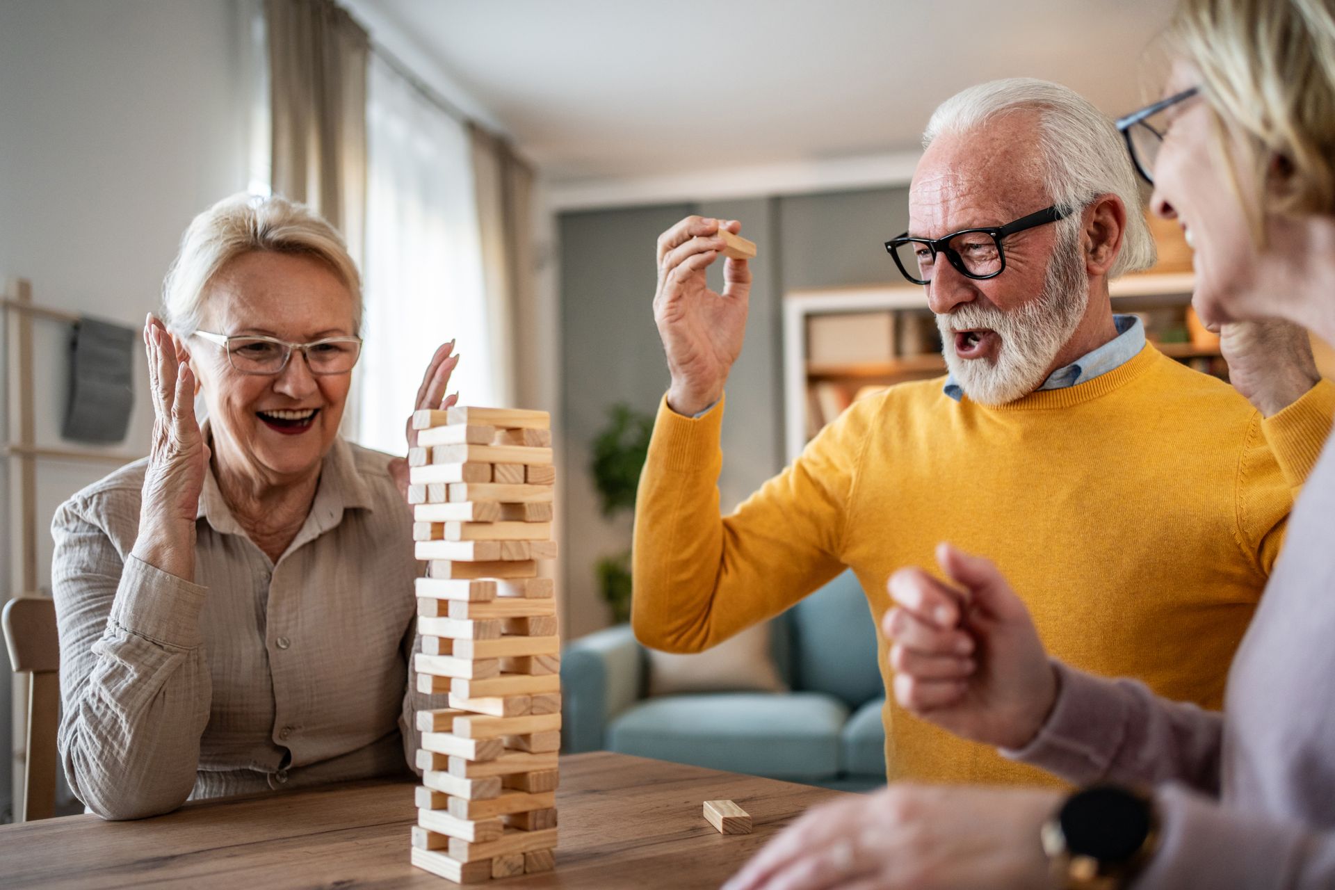 Three people play a tower-stacking game at a wooden table, laughing with excitement as the tower wobbles.