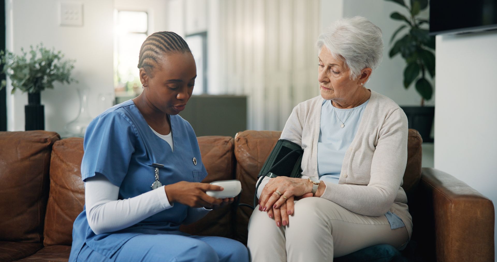 A healthcare worker in blue scrubs explains a medical device to a patient sitting on a leather sofa.