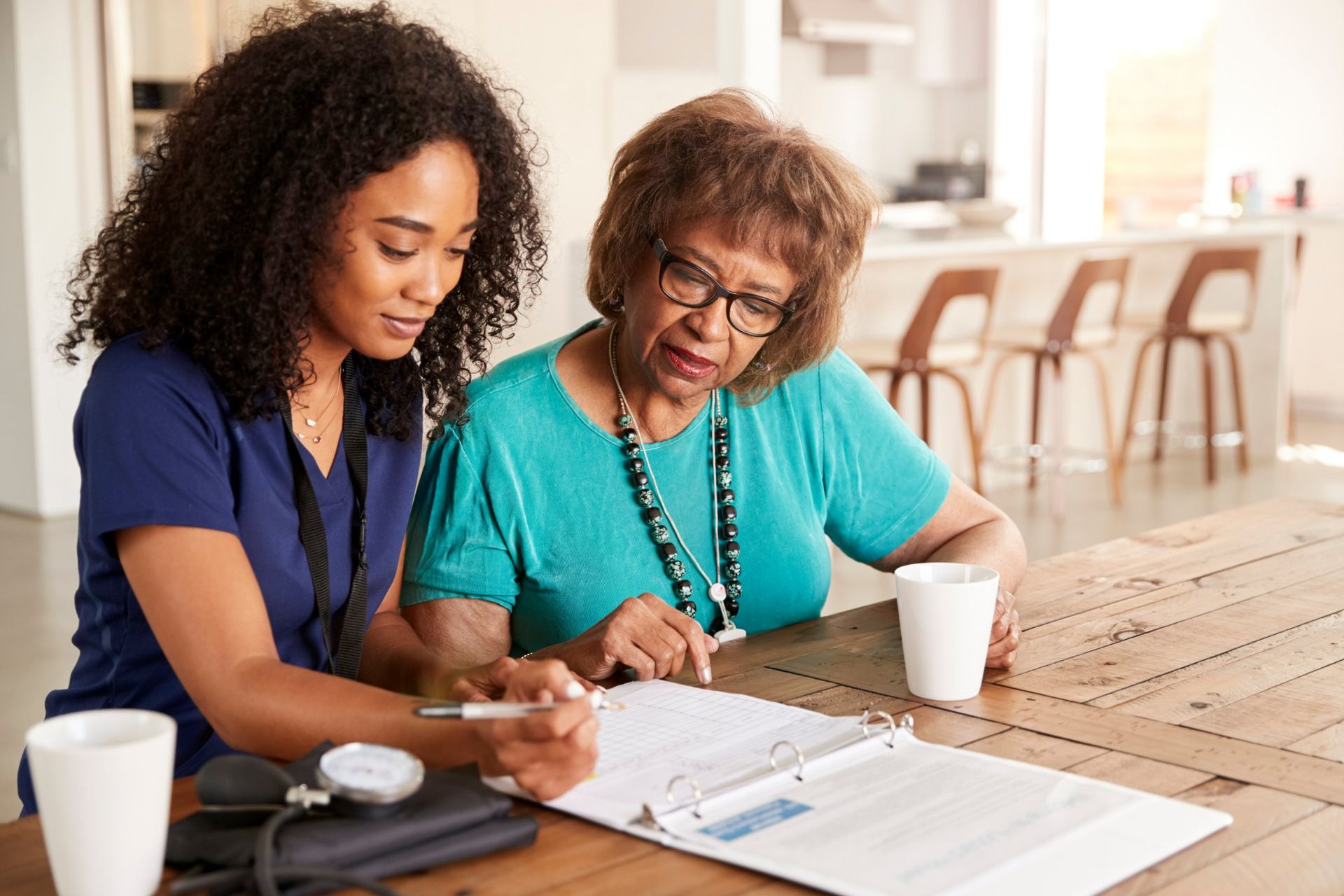 A healthcare worker in a blue uniform sits with a client at a wooden table, reviewing documents together.