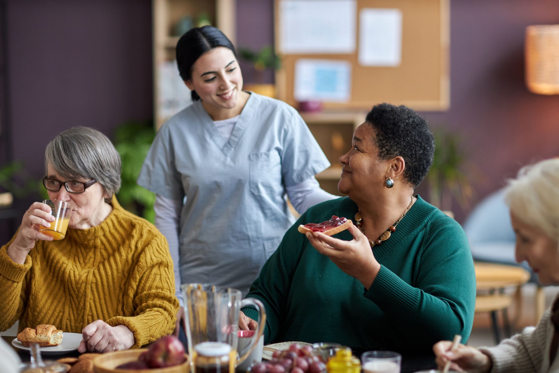 A staff member smiles while observing residents eating and drinking in a common area.