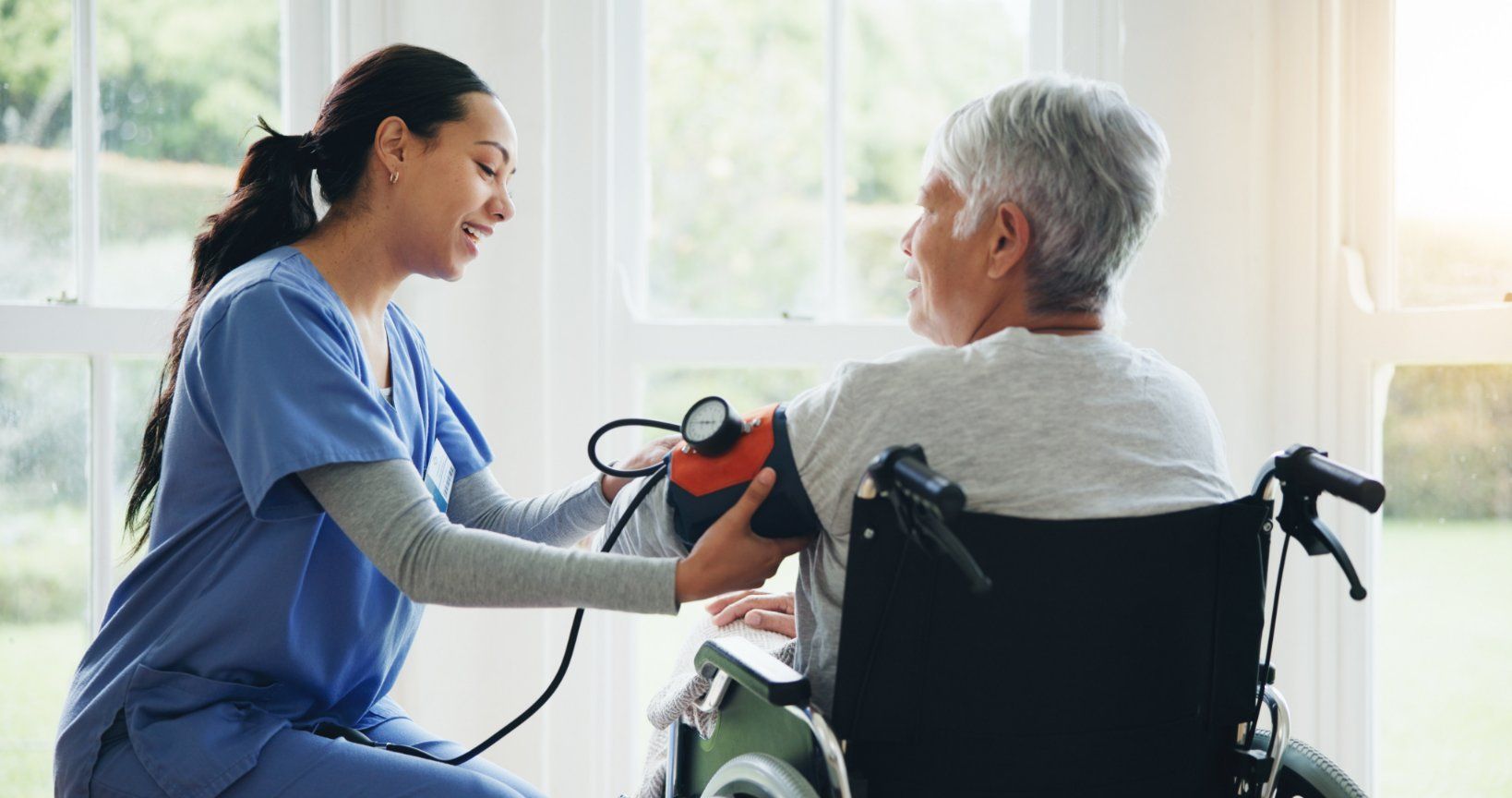 A healthcare worker in blue scrubs checks the blood pressure of a person sitting in a wheelchair near a large window.