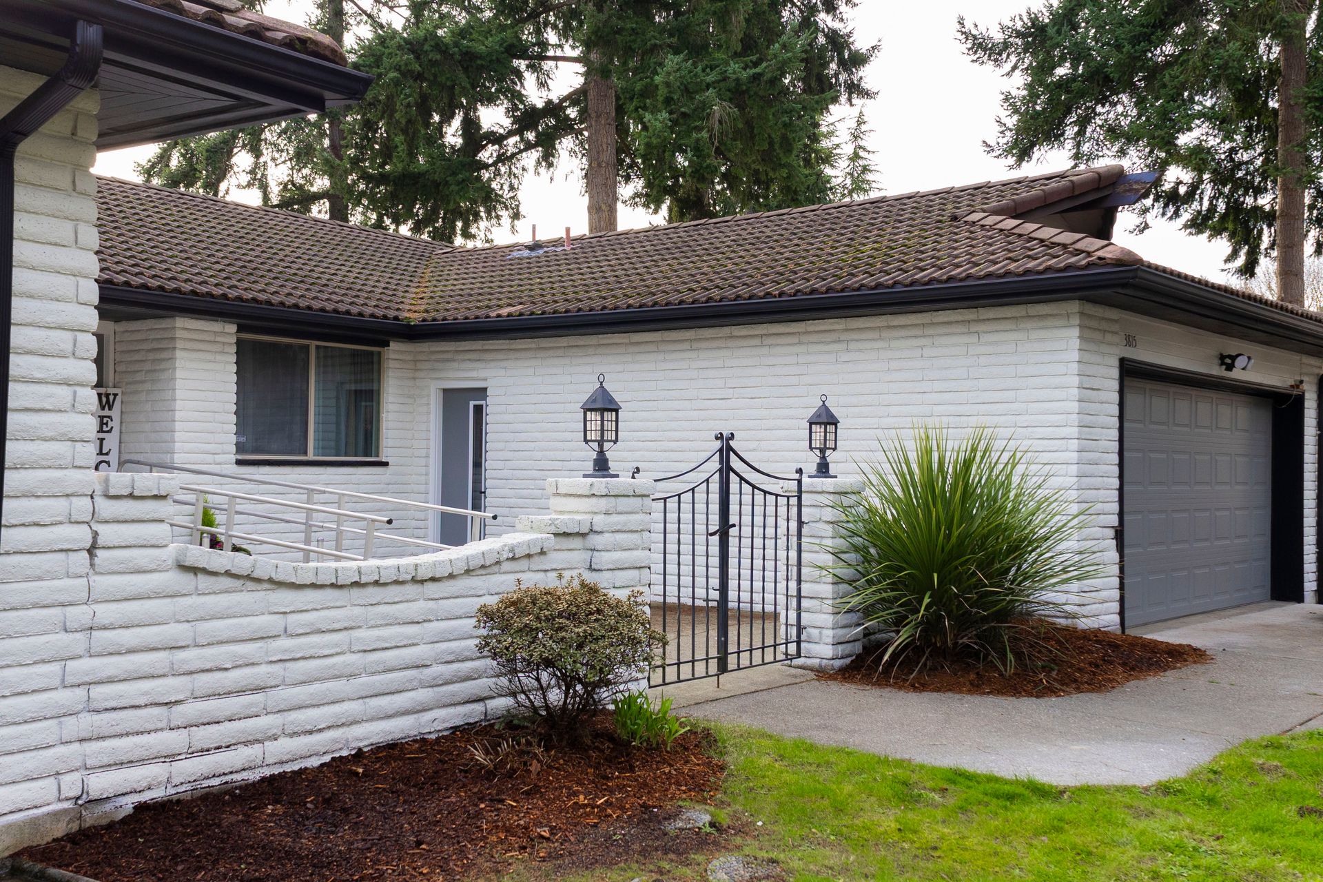A white-painted brick house with a brown shingled roof, a grey garage door, and a black wrought-iron gate in the front yard.