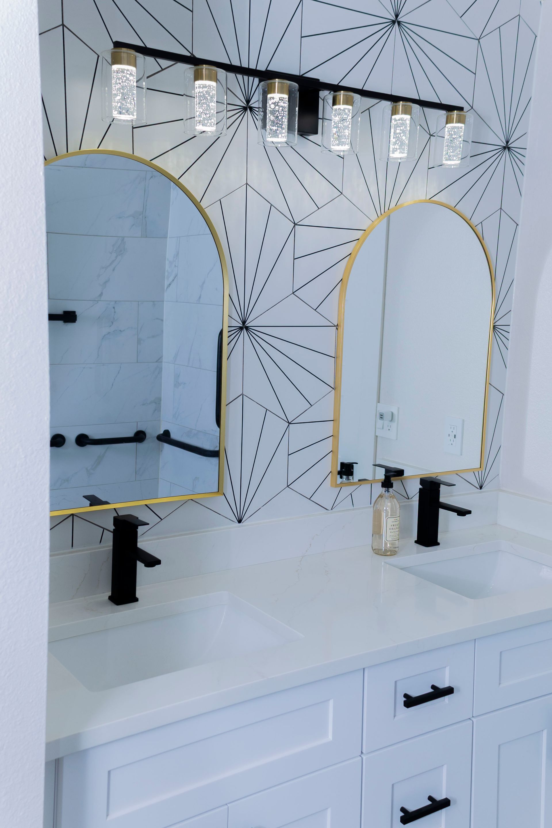 Modern double vanity with white cabinets, gold-rimmed arched mirrors, black faucets, and a geometric-patterned backsplash.
