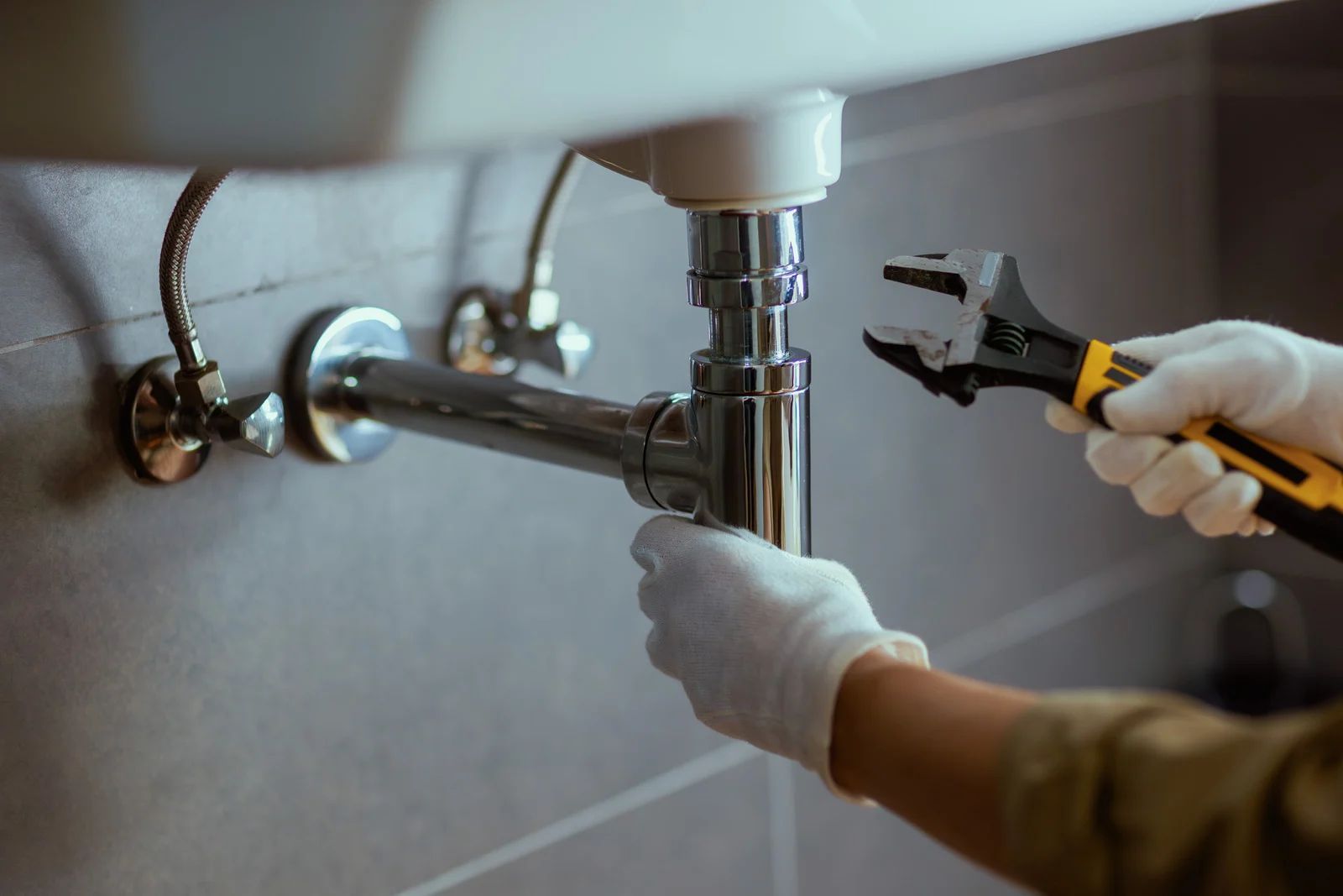 Plumber using wrench to work on sink pipes, wearing gloves.