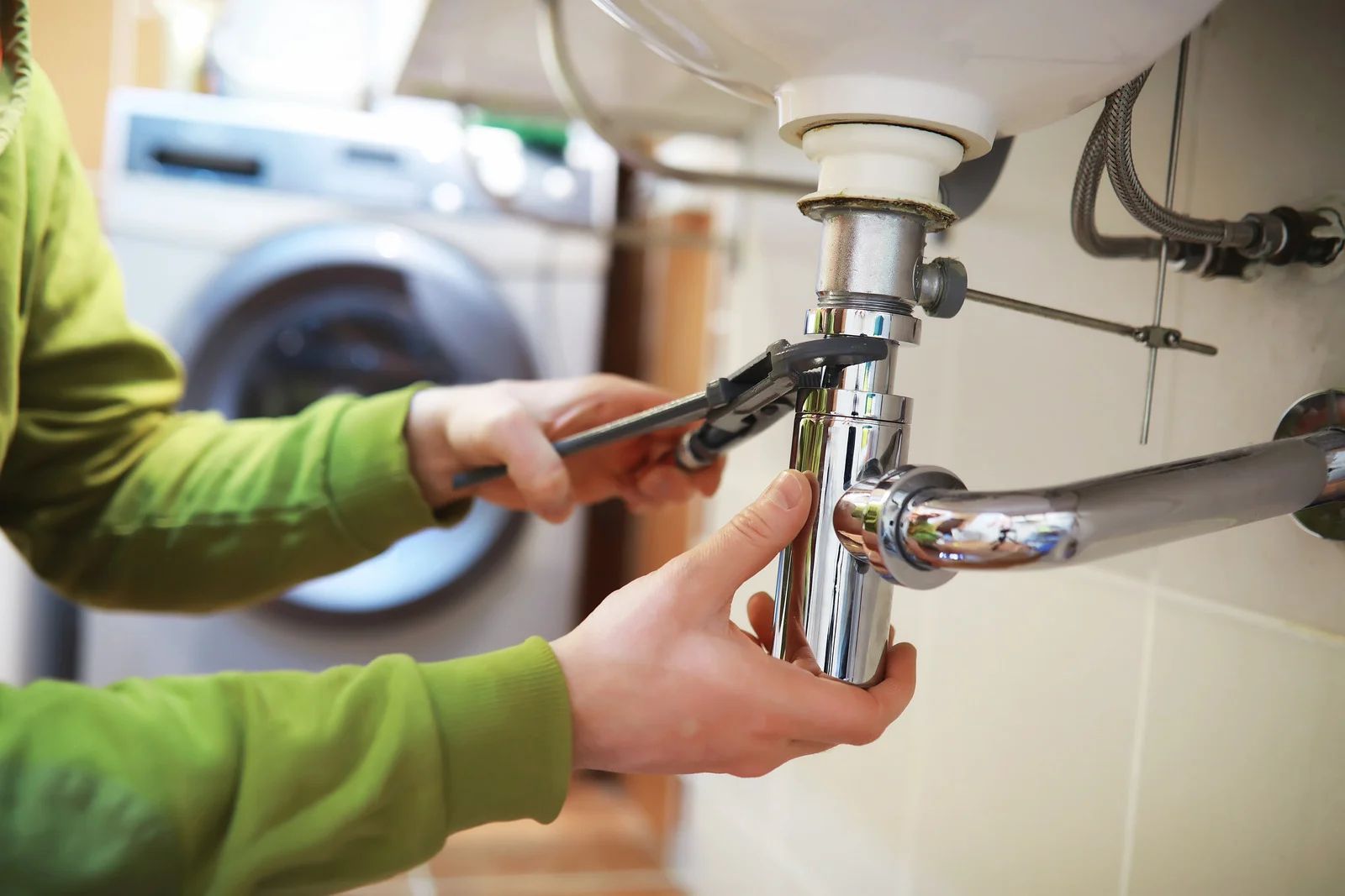 Person in green shirt using wrench to fix sink pipes. Laundry machine in background.