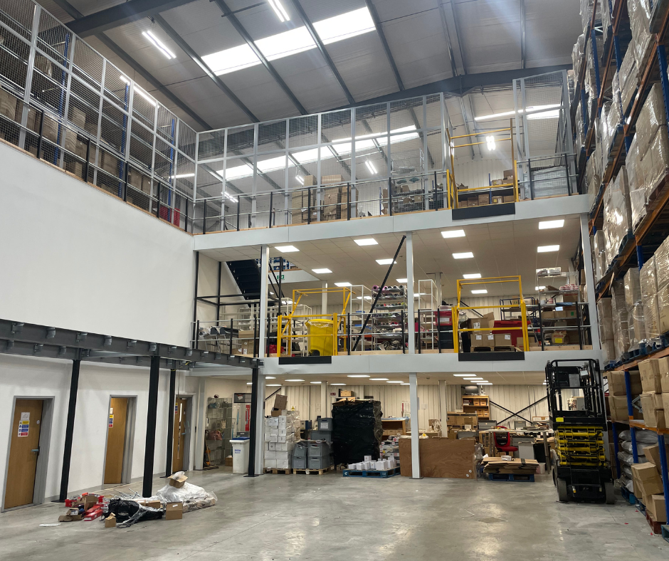Large warehouse interior with high shelves, mezzanine offices, pallets, and industrial equipment under bright lights