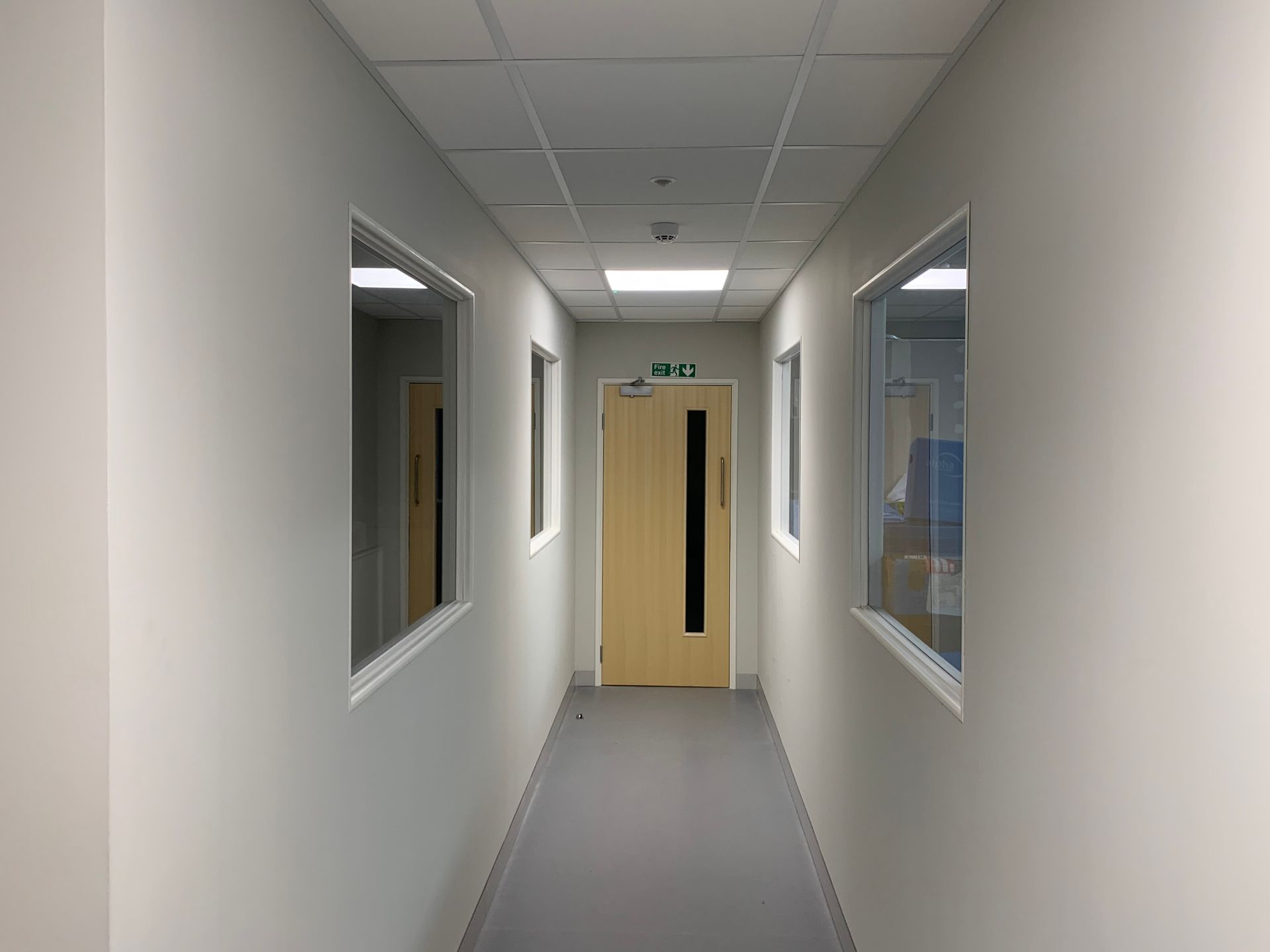 Empty office hallway with white walls, windows on both sides, and a light wood door at the end