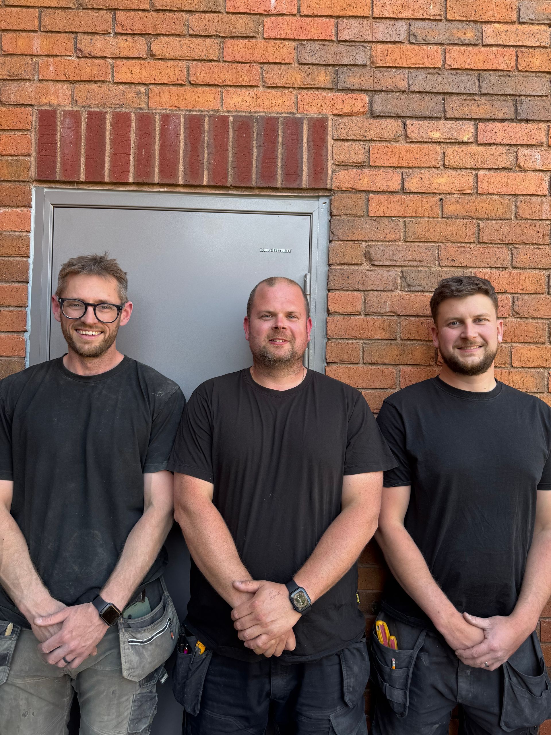 Three men in black shirts stand side by side in front of a brick wall and door.