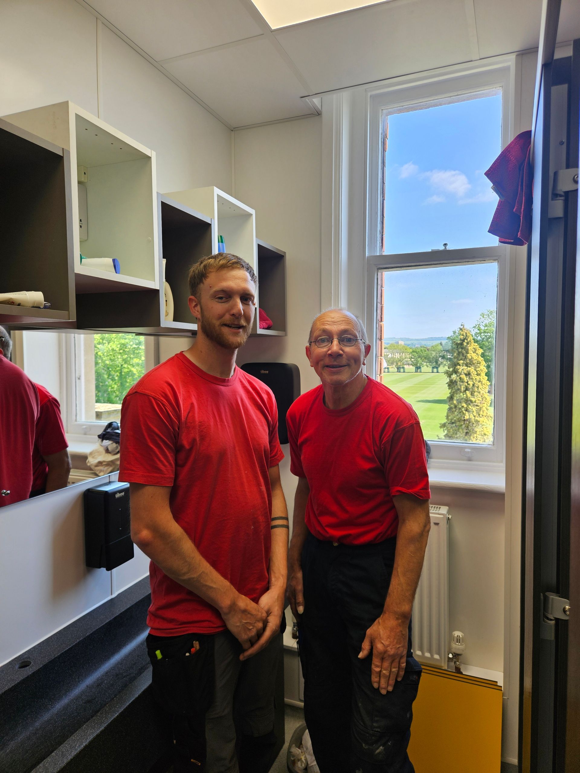 Two people in red shirts standing in a bright room beside open shelves and a window