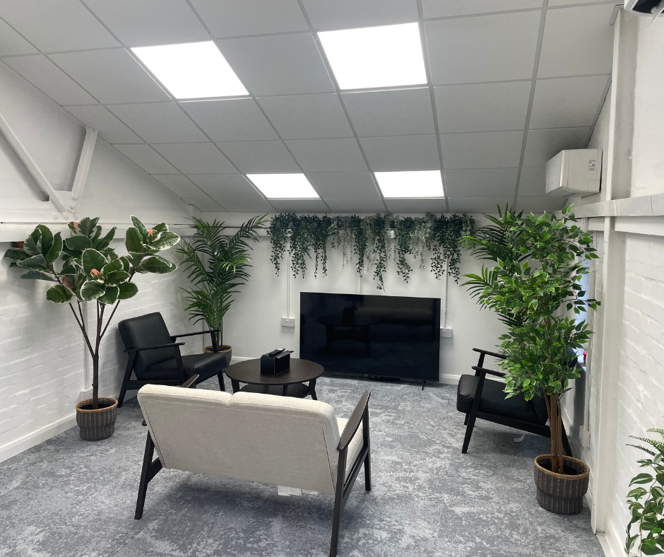 Modern lounge with gray seating, black chairs, potted trees, and a TV on a white wall