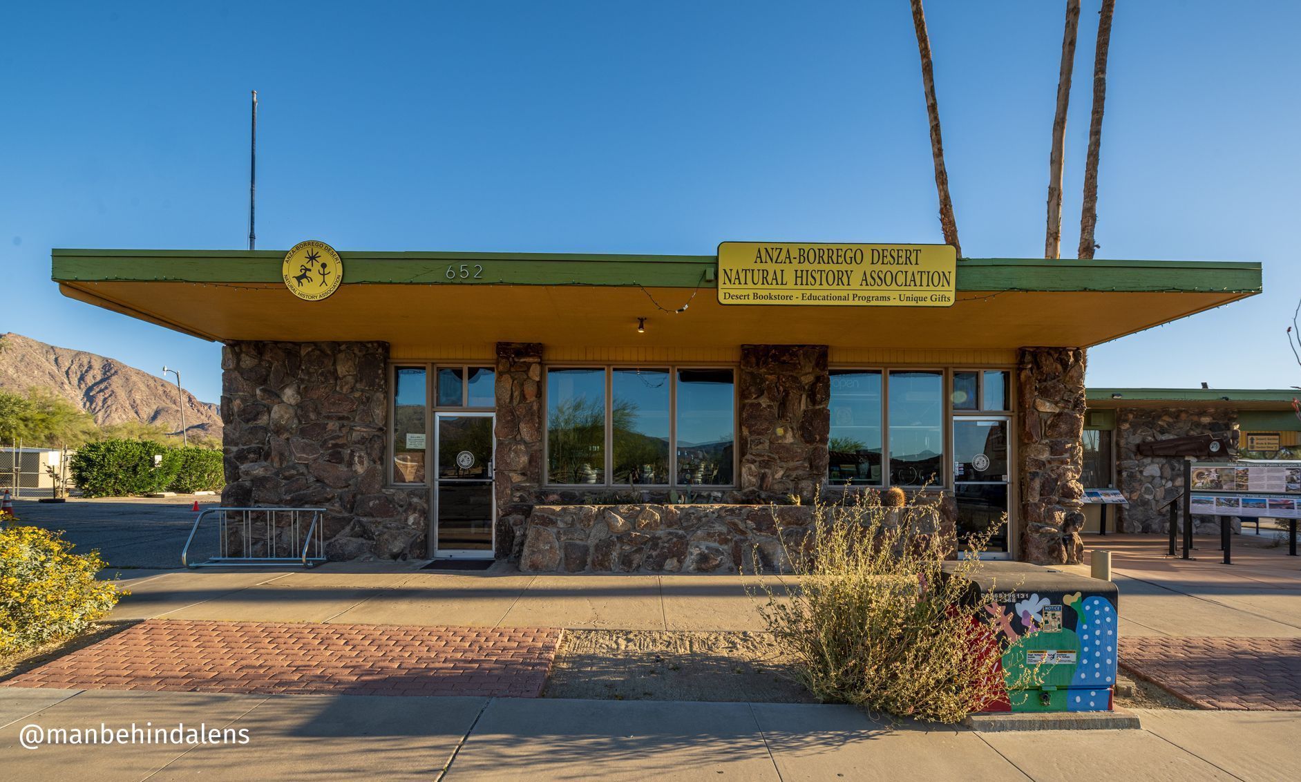 Small stone building with green roof, yellow sign, and desert landscaping under a clear blue sky