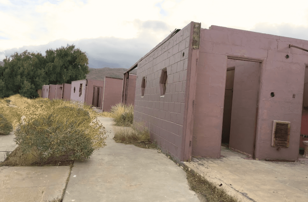 Adobe buildings along a desert path with a bush and cloudy mountains in the background